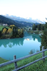 A picturesque landscape featuring a tranquil lake with turquoise water, surrounded by lush greenery and dense forest. In the background, majestic mountains tower under a clear sky, while a small village with quaint buildings rests at the base. A wooden fence runs along the foreground, adding a rustic charm to the scene.