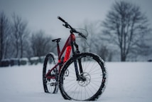 A red mountain bike is parked in a snowy landscape. Snowflakes are gently falling, and bare trees are visible in the background, creating a wintery scenery.