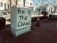 A metal utility box on a sidewalk has graffiti that reads 'Be <3 The Change.' The background includes several parked cars and residential buildings with trees that have no leaves lining the street.