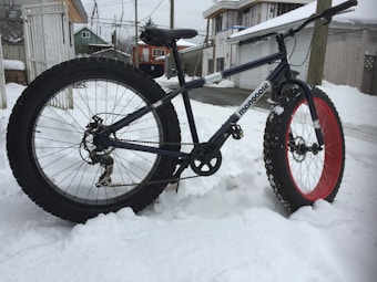 A fat-tire bicycle with one red rim and one black rim is positioned in a snowy environment. The background includes residential buildings and power lines, indicating an urban neighborhood.