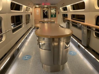 A modern train carriage with a sleek, curved countertop in the center. The interior features neutral tones with stainless steel elements, windows along the sides, and a clean, well-lit environment. There are indicators on the floor for social distancing and recycling bins integrated into the counters.