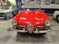 A classic red convertible car with a shiny exterior is prominently displayed in an indoor showroom. The car features chrome detailing and vintage design elements, including round headlights and a distinctive front grille. In the background, there are other cars and photos on the wall, suggesting a historical or museum setting.