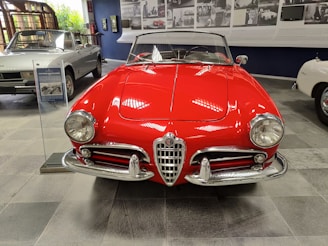 A classic red convertible car with a shiny exterior is prominently displayed in an indoor showroom. The car features chrome detailing and vintage design elements, including round headlights and a distinctive front grille. In the background, there are other cars and photos on the wall, suggesting a historical or museum setting.