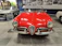 A classic red convertible car with a shiny exterior is prominently displayed in an indoor showroom. The car features chrome detailing and vintage design elements, including round headlights and a distinctive front grille. In the background, there are other cars and photos on the wall, suggesting a historical or museum setting.