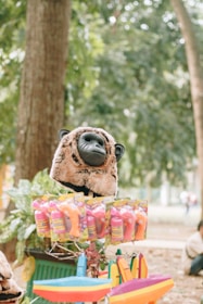 A stuffed animal head resembling a gorilla, displayed on top of a stall with colorful plastic toys and packaging. The background features lush green trees, giving a relaxed, natural setting.