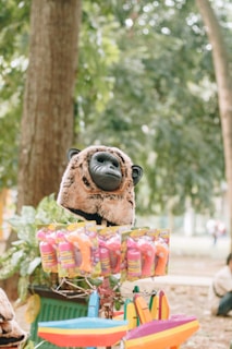 A stuffed animal head resembling a gorilla, displayed on top of a stall with colorful plastic toys and packaging. The background features lush green trees, giving a relaxed, natural setting.