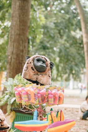 A stuffed animal head resembling a gorilla, displayed on top of a stall with colorful plastic toys and packaging. The background features lush green trees, giving a relaxed, natural setting.