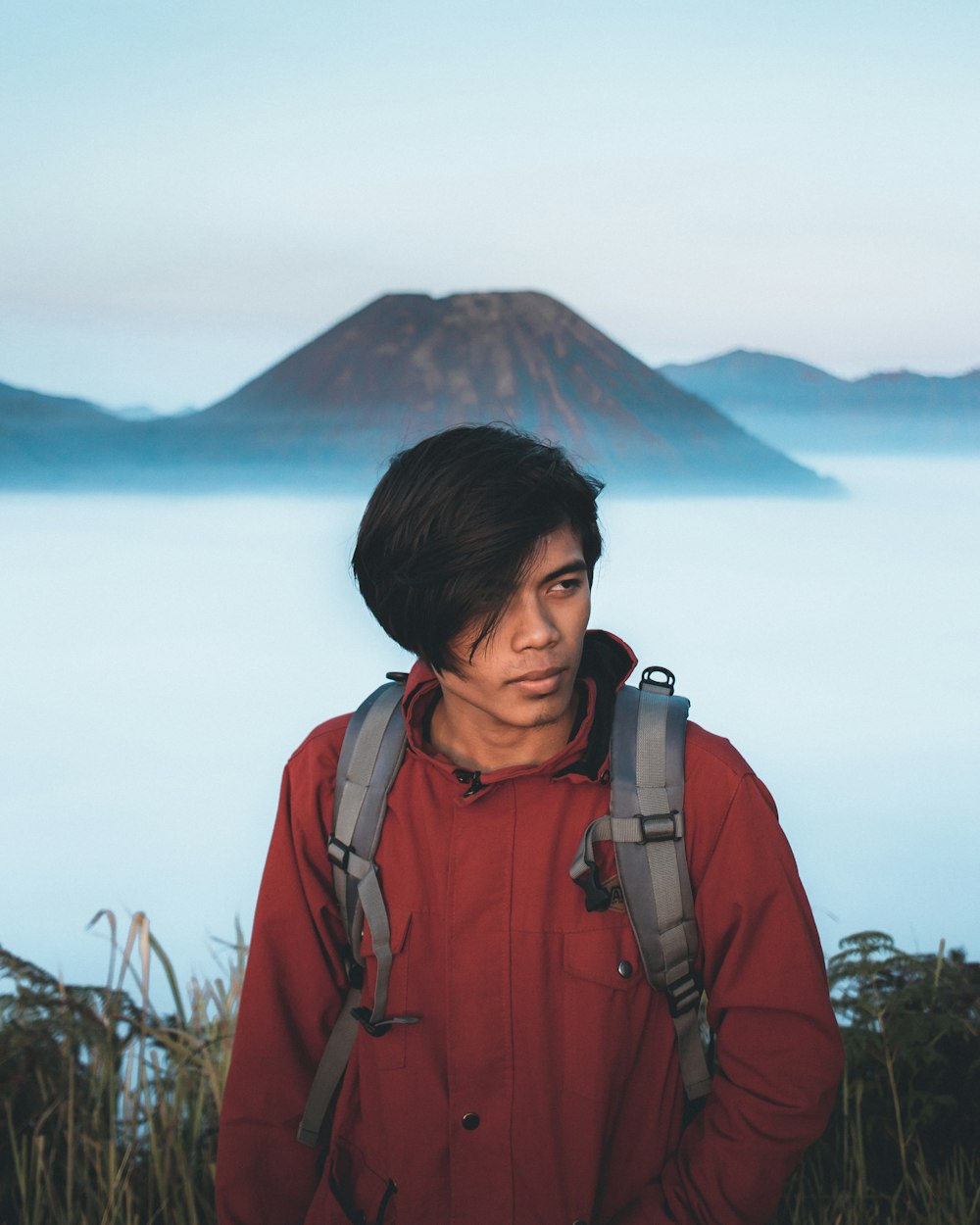 man in red zip up jacket standing near green trees and body of water during daytime