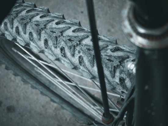 Close-up of a mountain bike tire with rugged tread patterns, showcasing the wheel spokes and axle. The tire and wheel appear worn and slightly dirty, emphasizing its use for off-road cycling.