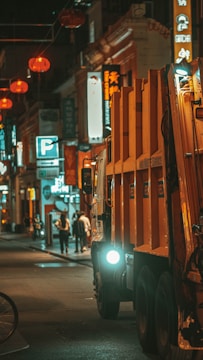 A nighttime urban street scene featuring a large orange waste collection truck in the foreground. The street is lined with various illuminated signs and lanterns, with a few people walking in the background. The setting appears to be in a busy city area with an array of commercial establishments.