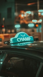 A taxi roof sign with the word 'CHAMP' is prominently displayed against a blurred urban nighttime background. The sign is illuminated and casts a reflection on the car roof, surrounded by bokeh lights in the background, creating a vibrant and dynamic scene.