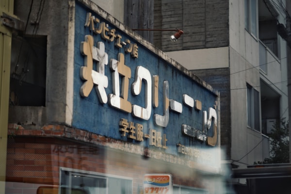 A weathered business sign with Japanese lettering is mounted on a building. The sign features a blue background with large white and yellow text. The building appears to be older, with some visible wear on the facade. There is a partly visible light fixture above the sign.