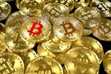 A large pile of shiny gold coins with the Bitcoin symbol embossed on them, with one coin featuring the symbol in red.