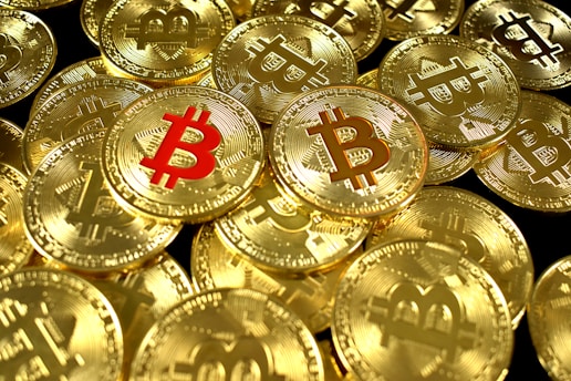 A large pile of shiny gold coins with the Bitcoin symbol embossed on them, with one coin featuring the symbol in red.