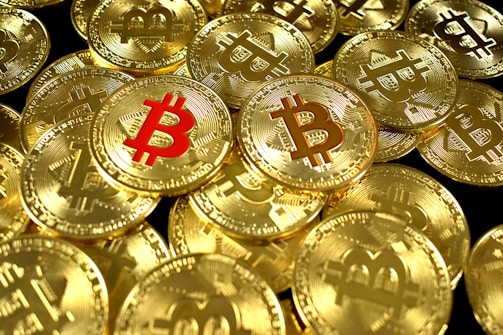 A large pile of shiny gold coins with the Bitcoin symbol embossed on them, with one coin featuring the symbol in red.
