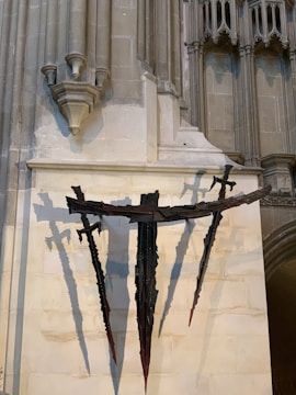 A stone wall with intricate gothic architectural elements. Dark, sculptural, cross-like figures with sword-like projections are mounted on the wall. They cast dramatic shadows against the light-colored stone, adding depth and a sense of mystery.