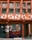 A traditional half-timbered building with an earthy color scheme of red and cream. The facade features large windows with green frames and a sign with the words 'Magasin Bio'. The lower part of the building displays window art with floral and plant motifs, incorporating orange and black tones.
