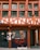 A traditional half-timbered building with an earthy color scheme of red and cream. The facade features large windows with green frames and a sign with the words 'Magasin Bio'. The lower part of the building displays window art with floral and plant motifs, incorporating orange and black tones.