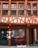 A traditional half-timbered building with an earthy color scheme of red and cream. The facade features large windows with green frames and a sign with the words 'Magasin Bio'. The lower part of the building displays window art with floral and plant motifs, incorporating orange and black tones.