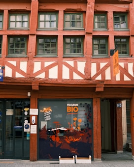 A traditional half-timbered building with an earthy color scheme of red and cream. The facade features large windows with green frames and a sign with the words 'Magasin Bio'. The lower part of the building displays window art with floral and plant motifs, incorporating orange and black tones.