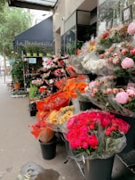 A charming outdoor flower shop displays an array of colorful bouquets. The florals include vibrant pink, red, orange, and white hues, neatly arranged in black buckets. A blue awning with lettering is visible above, and lush green plants are placed on shelves beside the shop entrance. The street and city life are subtly visible in the background.