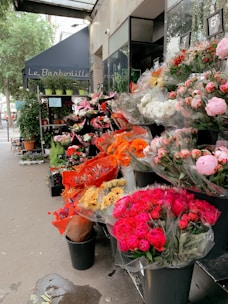 A charming outdoor flower shop displays an array of colorful bouquets. The florals include vibrant pink, red, orange, and white hues, neatly arranged in black buckets. A blue awning with lettering is visible above, and lush green plants are placed on shelves beside the shop entrance. The street and city life are subtly visible in the background.