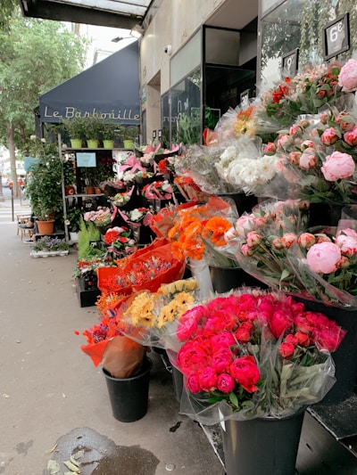 A charming outdoor flower shop displays an array of colorful bouquets. The florals include vibrant pink, red, orange, and white hues, neatly arranged in black buckets. A blue awning with lettering is visible above, and lush green plants are placed on shelves beside the shop entrance. The street and city life are subtly visible in the background.