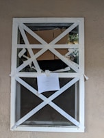 A closed window is secured with white tape arranged in an X pattern. Behind the window, there is cardboard blocking part of the view, and a paper note with the word 'CAUTION' is taped to the glass. The background wall is beige, and the overall setting appears to be an exterior scene.