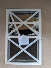 A closed window is secured with white tape arranged in an X pattern. Behind the window, there is cardboard blocking part of the view, and a paper note with the word 'CAUTION' is taped to the glass. The background wall is beige, and the overall setting appears to be an exterior scene.