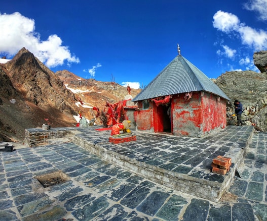 A small temple-like structure with a grey metallic roof and red-painted walls stands on a stone-tiled platform amidst a mountainous landscape. There are several red flags and some statues near the entrance of the building. In the background, rugged, partially snow-covered mountains stretch under a bright blue sky with scattered white clouds. A person is visible near the right side of the structure.