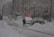 A snowy urban scene with several cars parked along a snow-covered street. A person is shoveling snow near the parked vehicles. Buildings line the street, and snow-covered trees are visible. Outdoor lamps and various street signs are also present.