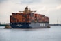 A large cargo ship laden with multicolored shipping containers is being assisted by two smaller tugboats in a wide expanse of water. The ship has the letters 'HMM' prominently displayed on its side. The background includes a distant industrial area with cranes and buildings.