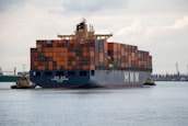 A large cargo ship laden with multicolored shipping containers is being assisted by two smaller tugboats in a wide expanse of water. The ship has the letters 'HMM' prominently displayed on its side. The background includes a distant industrial area with cranes and buildings.
