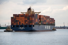 A large cargo ship laden with multicolored shipping containers is being assisted by two smaller tugboats in a wide expanse of water. The ship has the letters 'HMM' prominently displayed on its side. The background includes a distant industrial area with cranes and buildings.