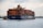 A large cargo ship laden with multicolored shipping containers is being assisted by two smaller tugboats in a wide expanse of water. The ship has the letters 'HMM' prominently displayed on its side. The background includes a distant industrial area with cranes and buildings.