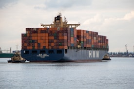 A large cargo ship laden with multicolored shipping containers is being assisted by two smaller tugboats in a wide expanse of water. The ship has the letters 'HMM' prominently displayed on its side. The background includes a distant industrial area with cranes and buildings.