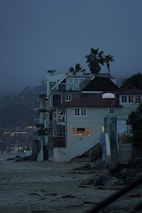 A row of beachside houses with multiple stories, featuring modern architecture with large windows showing indoor lighting. Palm trees rise above the structures against a misty, twilight sky. The sandy beach and rocky shoreline in the foreground create a contrast with the built environment.