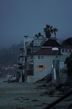 A row of beachside houses with multiple stories, featuring modern architecture with large windows showing indoor lighting. Palm trees rise above the structures against a misty, twilight sky. The sandy beach and rocky shoreline in the foreground create a contrast with the built environment.
