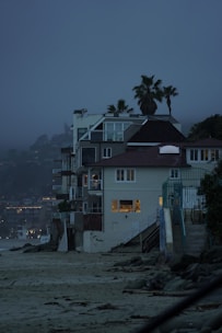 A row of beachside houses with multiple stories, featuring modern architecture with large windows showing indoor lighting. Palm trees rise above the structures against a misty, twilight sky. The sandy beach and rocky shoreline in the foreground create a contrast with the built environment.