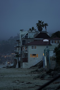 A row of beachside houses with multiple stories, featuring modern architecture with large windows showing indoor lighting. Palm trees rise above the structures against a misty, twilight sky. The sandy beach and rocky shoreline in the foreground create a contrast with the built environment.