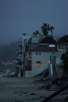 A row of beachside houses with multiple stories, featuring modern architecture with large windows showing indoor lighting. Palm trees rise above the structures against a misty, twilight sky. The sandy beach and rocky shoreline in the foreground create a contrast with the built environment.