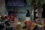 A small market stall, with a man in a red shirt organizing goods under a makeshift blue tarp roof. A woman wearing a hijab and patterned top stands nearby, engaging in conversation or purchasing. The area is cluttered with sacks of vegetables and goods, while a motorcycle with a red helmet is parked in the foreground. A tree trunk is partially visible on the right side.