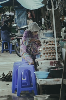 A person wearing a floral-patterned garment and a face mask is seated at a small outdoor market stall. They are handling eggs, which are stacked in cardboard cartons on the table. Nearby are blue plastic stools and a motorcycle parked beside the stall. The scene is shaded by makeshift covers.