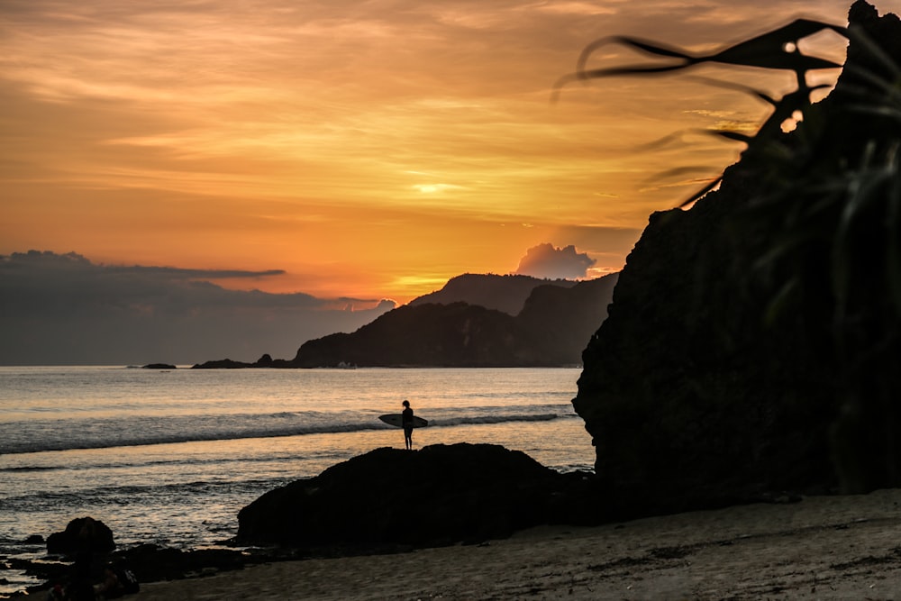 silhouette of person standing on rock near body of water during sunset