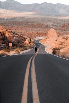 A winding desert highway stretching into a sunset sky, with a lone figure walking beside a vintage motorcycle.
