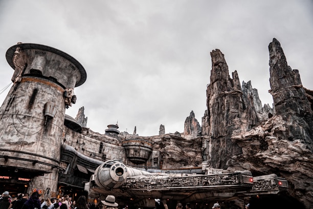 A futuristic setting with a large, iconic spacecraft positioned in front of towering rock formations that resemble a spaceport or otherworldly cityscape. The environment is bustling with visitors, some wearing hats, against a cloudy sky.