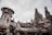 A futuristic setting with a large, iconic spacecraft positioned in front of towering rock formations that resemble a spaceport or otherworldly cityscape. The environment is bustling with visitors, some wearing hats, against a cloudy sky.