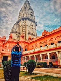 A person stands in a prayer pose facing a large, intricately designed temple with tall spires. The temple features architectural details in shades of pink and gray, set against a partly cloudy sky. Manicured bushes and a well-maintained courtyard surround the foreground.