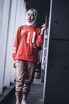 A person wearing a white hijab and a red long-sleeve shirt with print graphics stands leaning against a wall. The person also wears light brown jogger pants and white sneakers. The setting appears to be indoors with natural light coming from large windows.