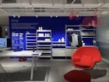 A modern interior space showcasing a minimalist shelving system against a vibrant blue wall. The shelves hold decorative items such as white vases and folded garments. A desk with a computer is positioned on the left side, while a bright red chair and ottoman set add a splash of color to the room. Industrial lighting fixtures hang from the ceiling.
